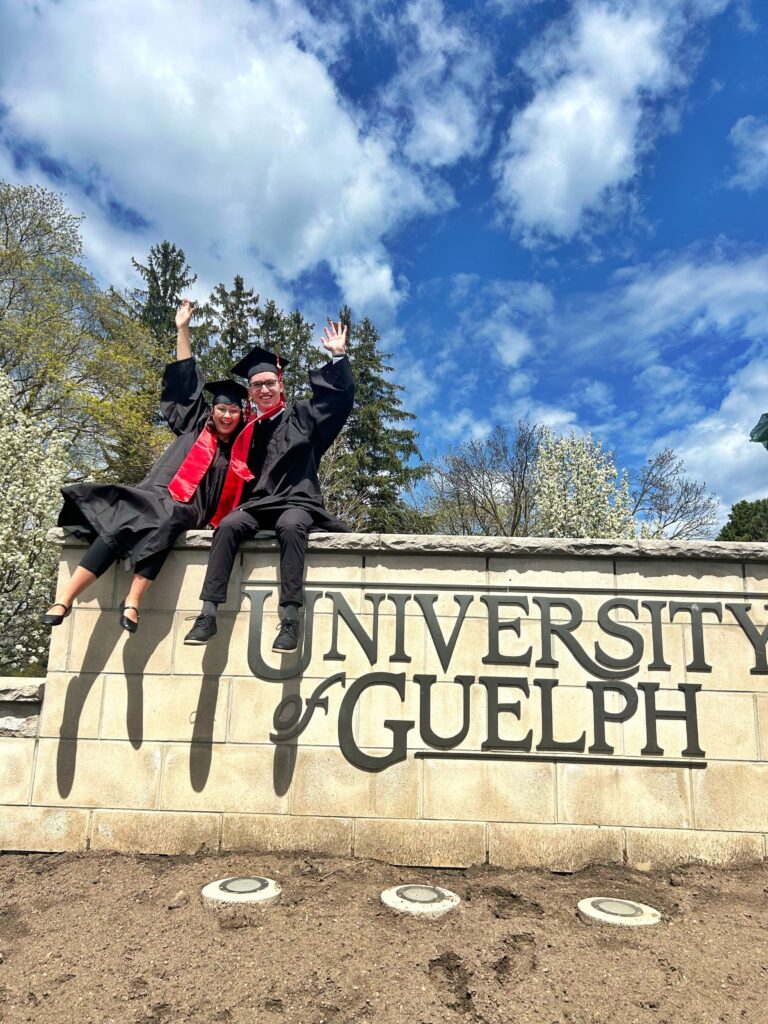 Matia and Mackenzie celebrating after their graduation. The two Campus Friends graduates are dressed in grad gowns and caps are sitting on top of the University of Guelph sign