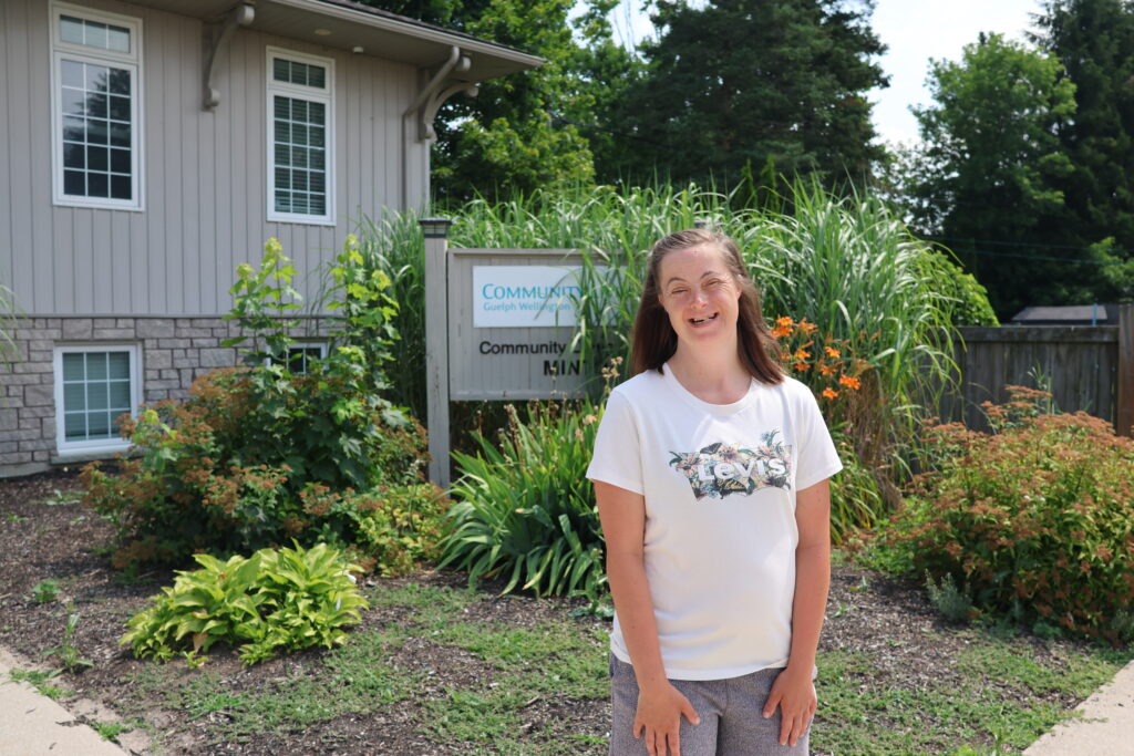 A woman with a developmental disability is standing in front of the Community Living Guelph Wellington sign and building