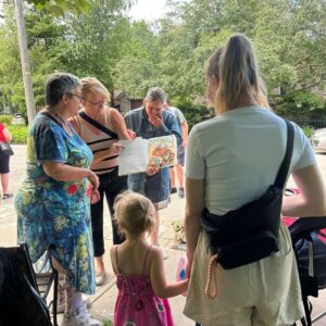 A woman holds open a book among a group of people at the Accessible Little Library